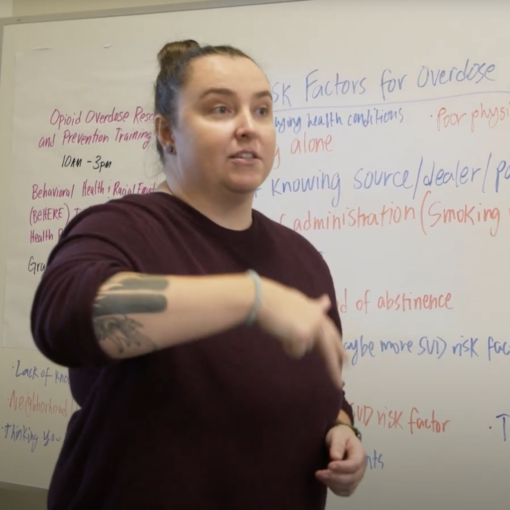 White woman in maroon sweater standing in front of a board with some writing on it about opioid overdose prevention.